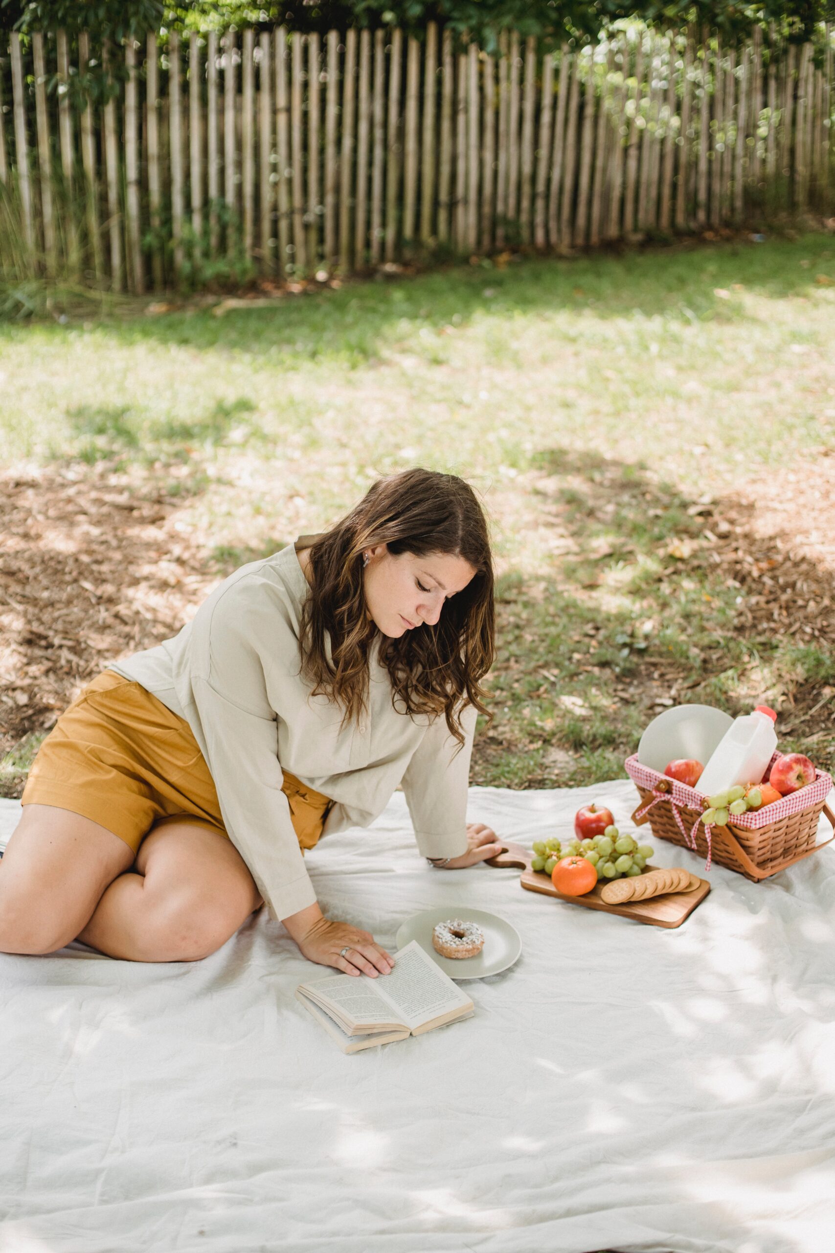 Pregnant woman in white top and yellow shorts sitting on picnic blanket reading a book with bagel and fruit.